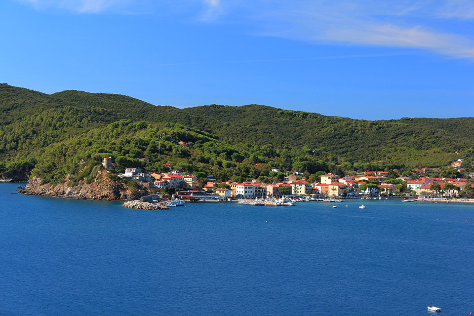 Blick vom Wasser auf das Küstendorf Marina di Campo auf Elba, umgeben von bewaldeten Hügeln