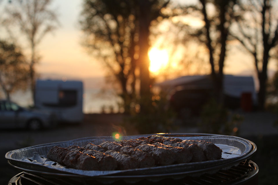 Gegrilltes Fleisch auf dem Campinggrill im Abendlicht, im Hintergrund der Camper