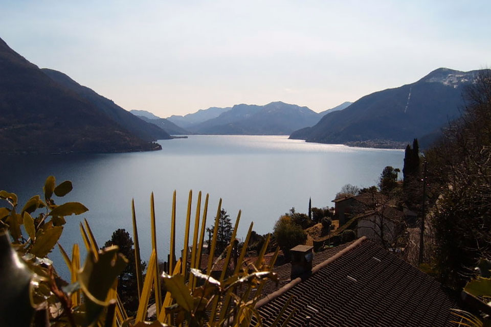 Blick über den Lago Maggiore auf die umliegenden Berge, im Vordergrund ein Dach mit Kamin