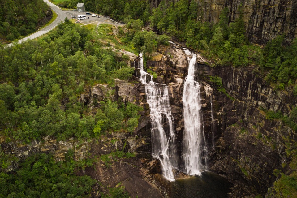 Doppelter Wasserfall stürzt eine bewaldete Felswand hinunter, direkt neben einer Passstrasse