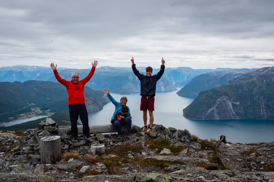 Drei Personen strecken auf einem Aussichtspunkt jubelnd die Arme in die Höhe, im Hintergrund ein Fjord
