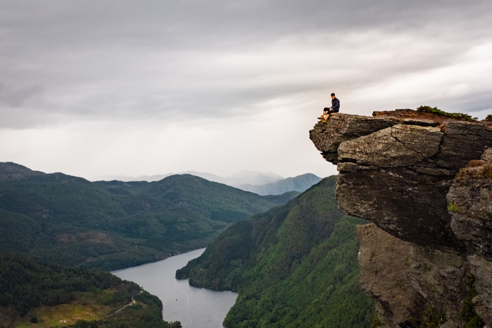 Person sitzt auf einem überhängenden Felsvorsprung mit Blick auf einen bewaldeten Fjord