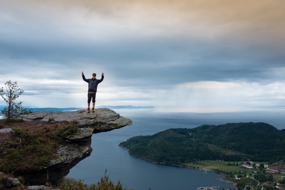 Person steht mit erhobenen Armen auf einem Felsvorsprung über einem Fjord