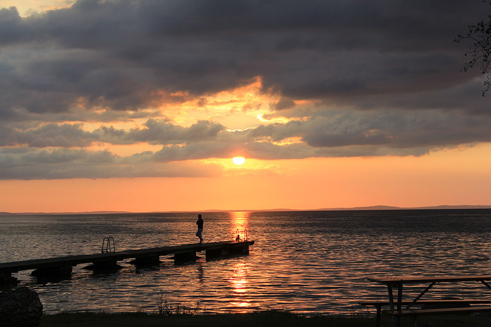 Person auf einem Holzsteg beobachtet den Sonnenuntergang über dem Vätternsee