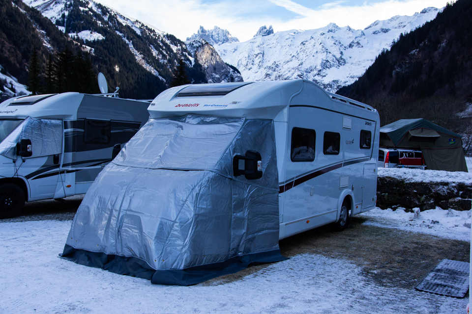 Camper mit Isolierhaube vor dem Fahrerhaus auf dem winterlichen Campingplatz, Berge im Hintergrund