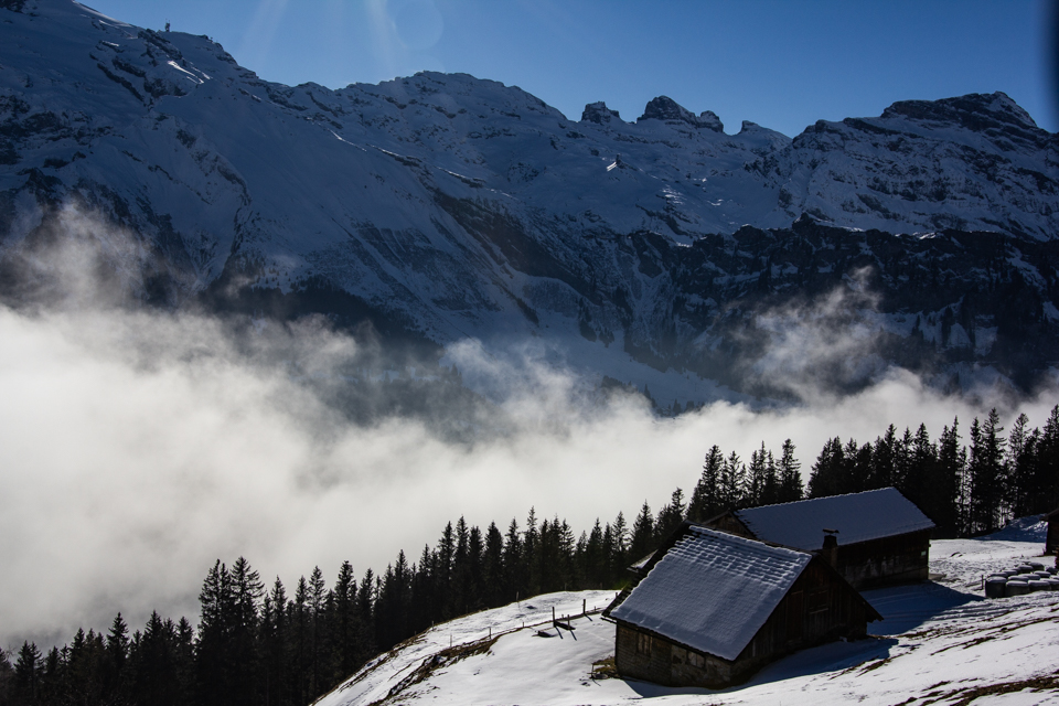 Alphütte über einem Nebelmeer, dahinter schneebedeckte Berggipfel